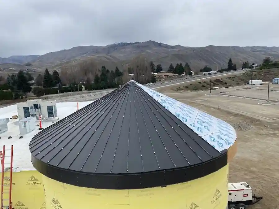 Black conical standing-seam roof on a round building under construction, mountains behind